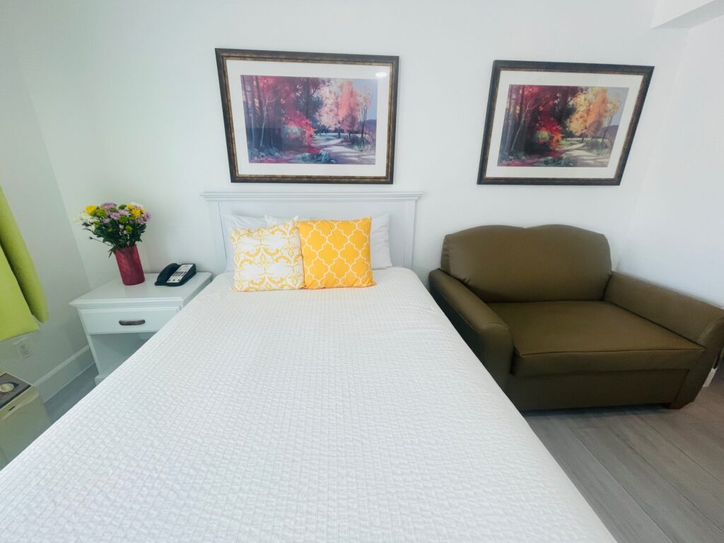 Size full bed with white linens and yellow accent pillows in a hotel room with two paintings on the wall above.  Brown leatherette folding sofa bed is on the right, and a small nightstand with a vase of flowers is on the left.