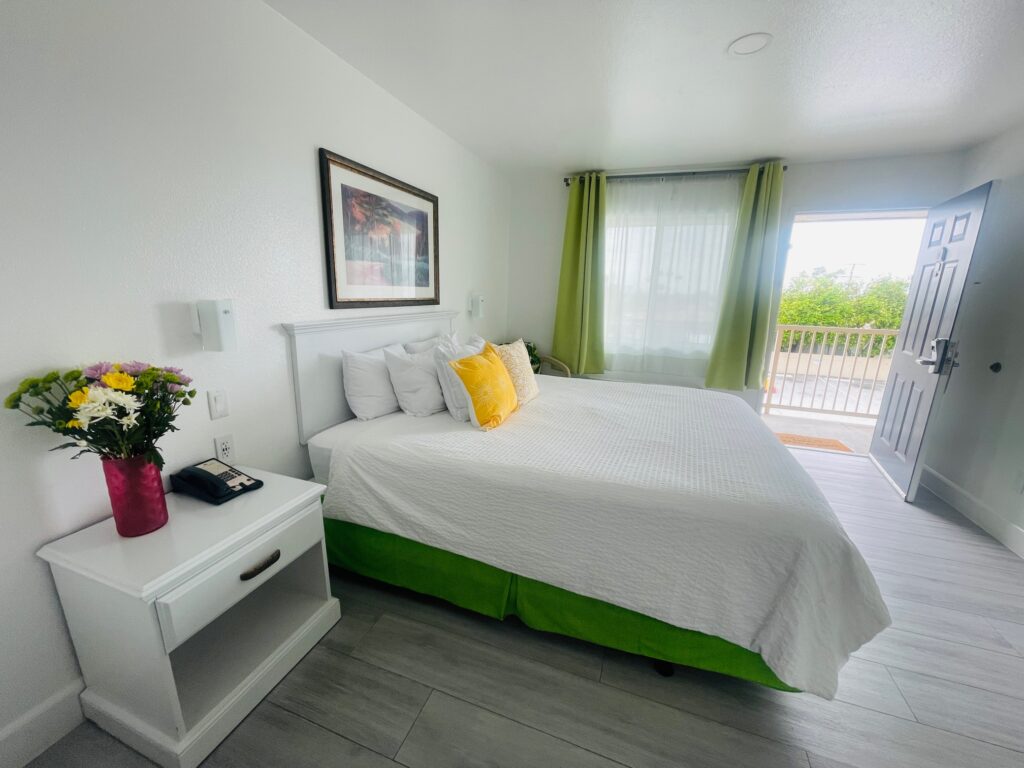 White Cal-king size bed in a bright hotel room.  An open door is on the right, and a nightstand with a phone and flower display is on the left.